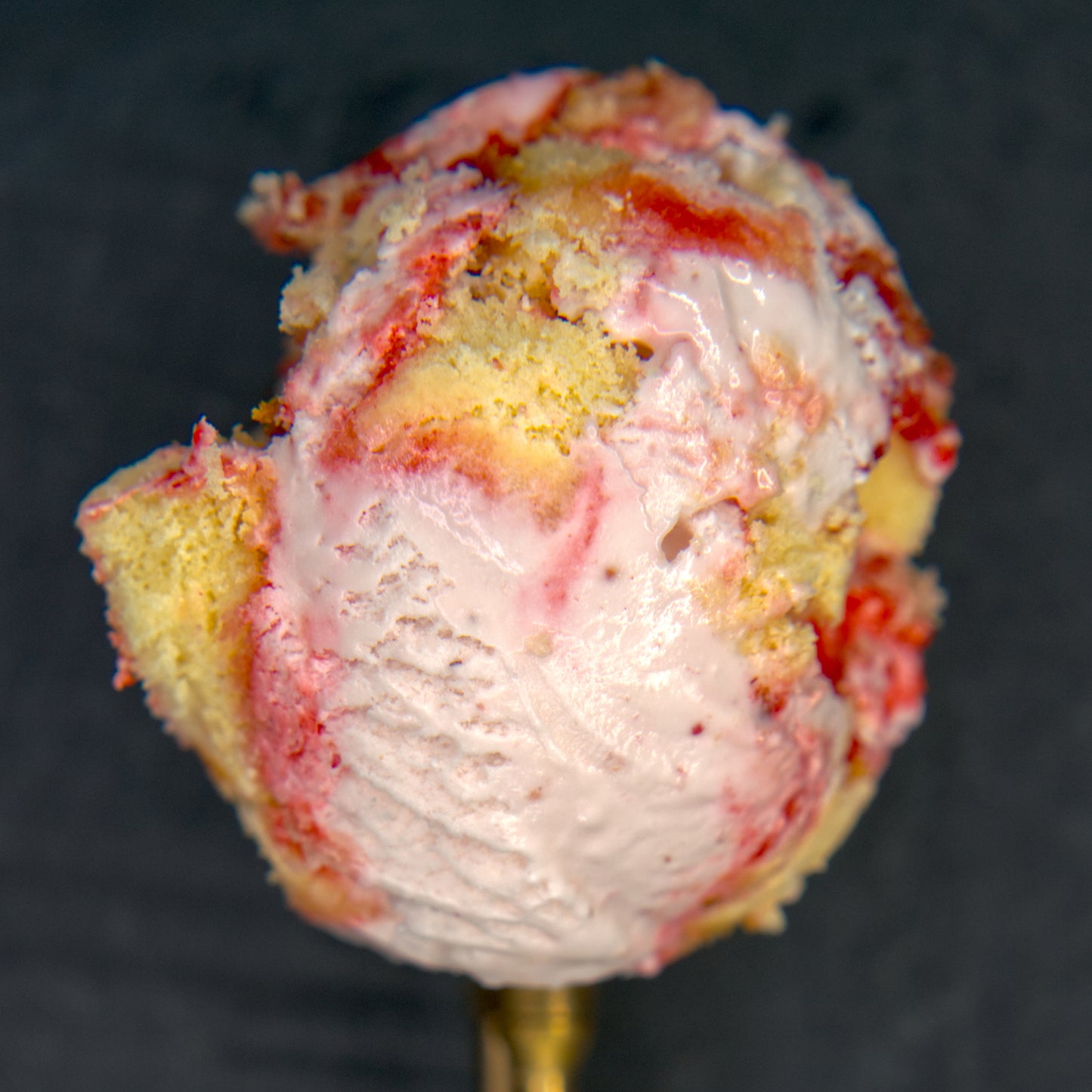 Ice cream scoop with a strawberry swirl and piece of cake against a dark background.