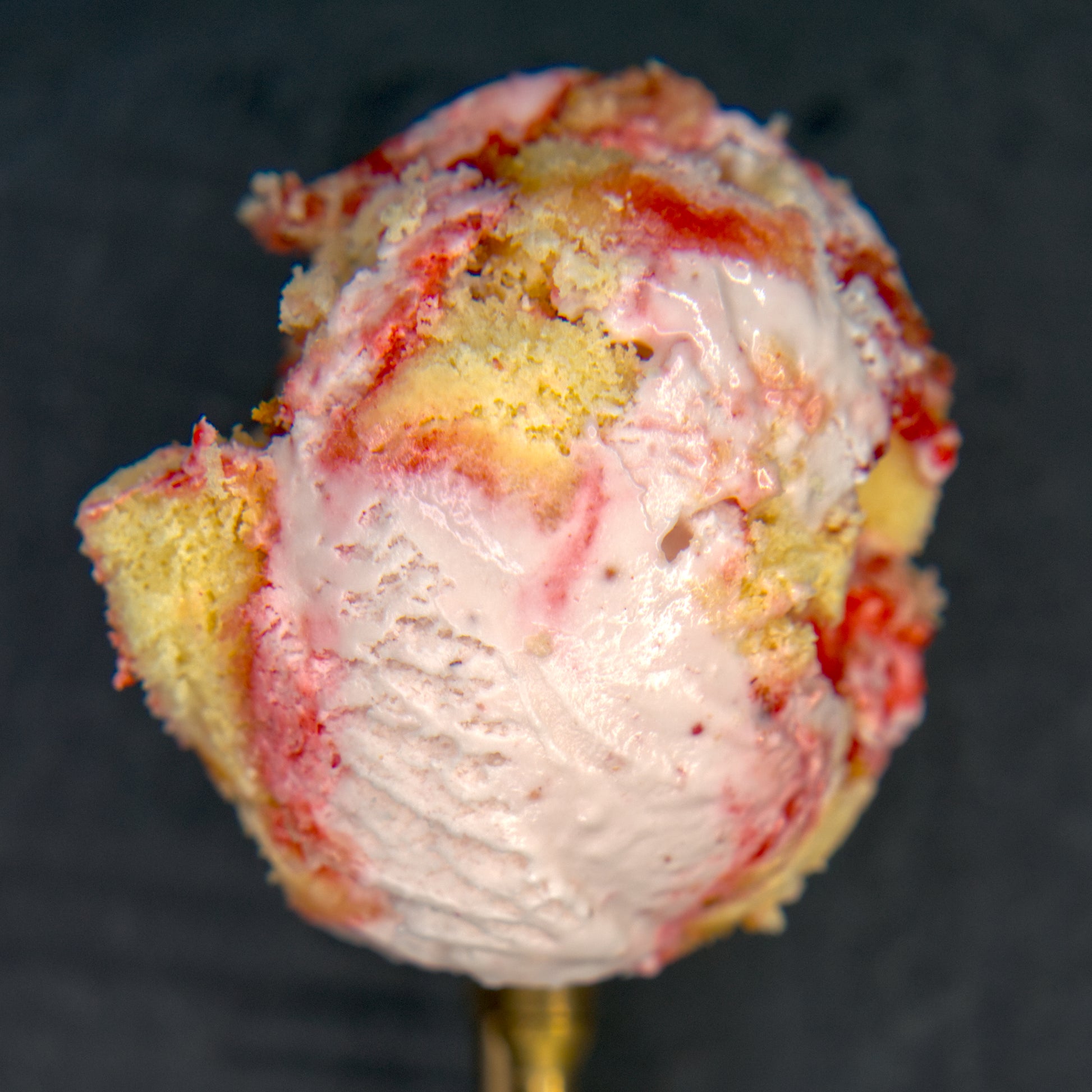 Ice cream scoop with a strawberry swirl and piece of cake against a dark background.